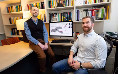 Two professors, one sitting on a desk with a black vest and blue collared shirt, the other seated at a computer, look facing the camera as a graphic display of AI in gaming is shown on an Apple iMac computer with a row of books behind the computer.