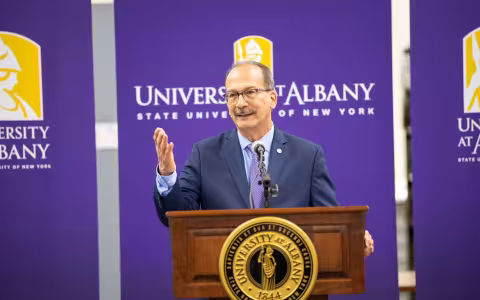 President Rodriguez stands behind a podium in front of UAlbany banners