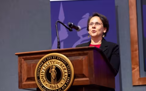 A woman in a black blazer stands behind a brown lectern with a gold University at Albany seal on the front, speaking into a microphone.