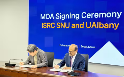 Two men sit a a brown desk signing documents in front of a blue screen that says "MOA Signing Ceremony"