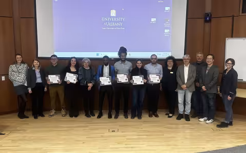 Fourteen people pose for a group portrait in front of a pull-down projection screen displaying the UAlbany logo. All are smiling and six are holding white paper certificates.