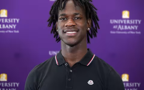 An African Americna young man wears a black collared shirt while standing against a purple backdrop with University at Albany written on it.