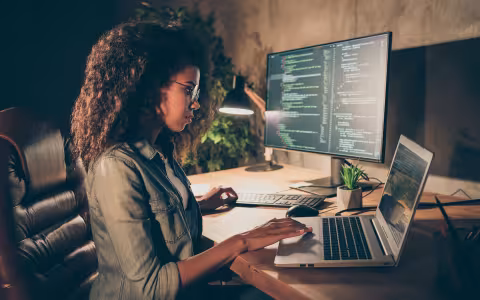 A cybersecurity professional works on a laptop alongside a monitor displaying code.