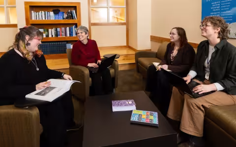 Four seated people smile in lively conversation. Two are using laptops, and the two others are holding a book and a notebook.