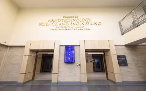 A sign for the College of Nanotechnology, Science, and Engineering hangs above two doorways in a building with light tan and brick walls.