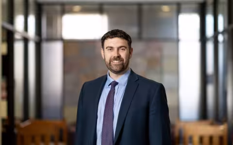 A smiling portrait of a bearded man with a dark blue suit and purple tie. 