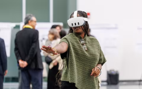 A woman with long braids and a green sweater wears a virtual reality headset and gestures in front of her.