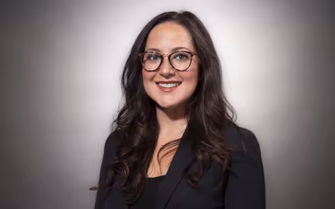 A woman with long dark hair and glasses in a black blazer smiles for a portrait against a gray backdrop.