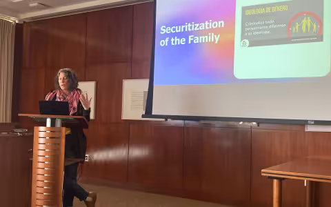 An image of Dr. Annie Wilkinson standing in front of a podium, a drop-down screen behind her with a "Securization of the Family" slide.