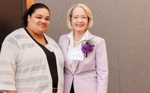Two smiling women stand together wearing nametags. The older woman has flowers in her lapel.