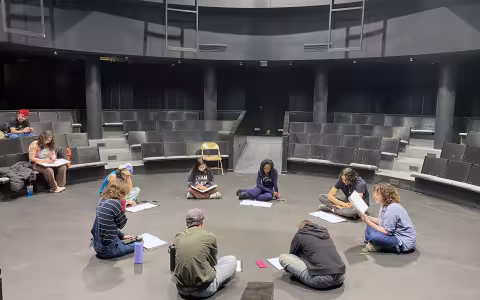 A group of students sit in a theatre-in-the-round with dark seats and walls as they sit in a circle with scripts in front of them.