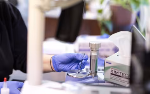 Using a small metal instrument, a person wearing purple latex gloves arranges a tiny amount of a white substance in the center of a round silver platform attached to a piece of lab equipment.
