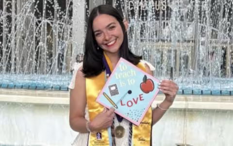 A young woman with shoulder-length dark hair wears a golden sash and holds a graduation cap in front of the UAlbany fountain that reads "To teach is to love"