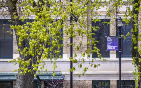 Tree leaves in spring bloom frame a brick building and lamppost bearing a purple banner that reads "College of Nanotechnology, Science, and Engineering" below the Minerva logo.