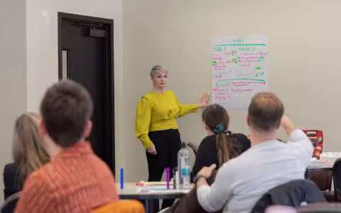 An instructor speaks to a classroom full of students, pointing to a large piece of paper with information on the wall.