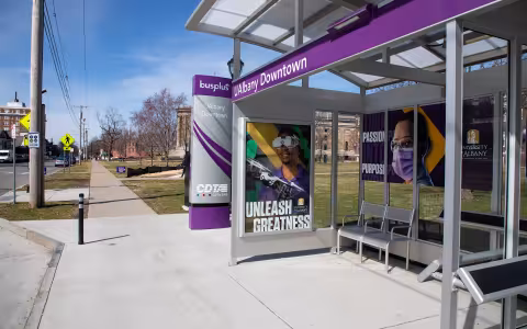 A purple CDTA bus shelter displaying “UAlbany Downtown” and “Unleash Greatness” signage, with metal seating beneath the covered glass structure.