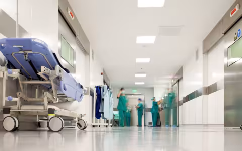 Healthcare workers wearing green scrubs, masks and surgical caps work at the end of a brightly lit hospital hallway.