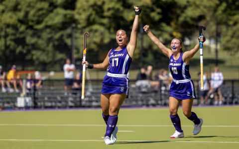 A young woman with dark brown hair in a purple field hockey uniform of the UAlbany Great Danes wearing number 17 lifts her arm in celebration on the turf field.