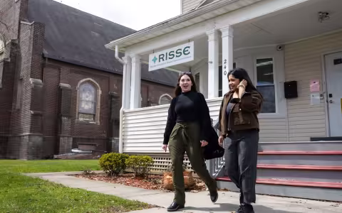 Two women smile as they exit a building with a sign reading: “RISSE Refugee & Immigrant Support Services of Emmaus” hanging from the porch roof. A brick church is visible in the background. 