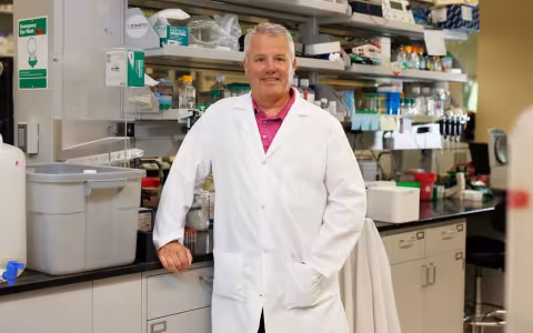 A man wearing a white lab coat and salmon shirt smiles for a portrait in a biology lab.