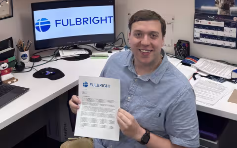 Chris Gilberti holds up a Fulbright offer in front of a computer screen.