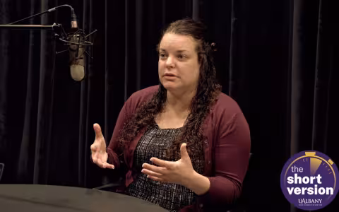 A woman in a maroon sweater sits in front of a microphone against a black background while she gestures with her hand. The words "the short version" are in a purple circle in the lower righthand corner.