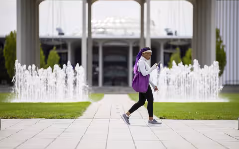 A UAlbany student walks across campus while looking at her phone, with a fountain and the Academic Podium in the background.