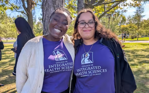 A portrait of two students wearing purple College of Integrated Health Sciences Shirts. They are outside, with trees and grass behind them.