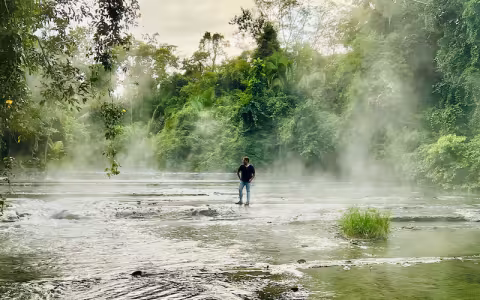 A man stands on a rock in a steaming river flowing through a jungle.