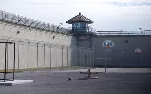 an empty prison yard surrounded by chain-link feancing and a high wall, both topped with razor wire.