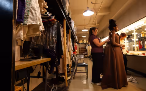 A woman stands in a dressing room with bright lights while costumers help fit her for her dress.