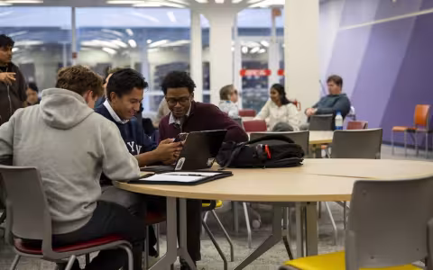 Smiling students seated at a table huddle over a laptop and a phone 