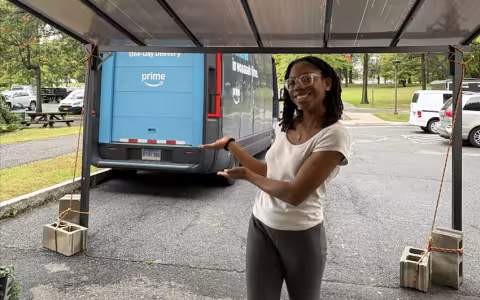 A student in a white top and gray slacks smiles and gestures toward an Amazon delivery truck