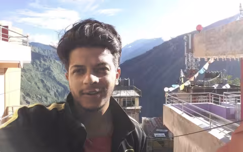 A young man takes a selfie on a balcony with a view of a mountain valley behind him.