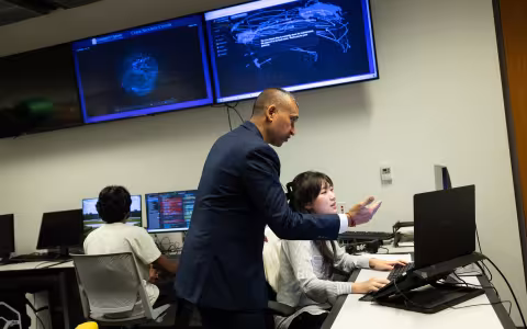 A professor stands by a female student at a computer as he gestures toward her screen and another student sits in the background.