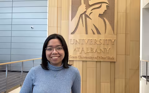 Shyrill Mariano smiles in front of a sign for the University at Albany.