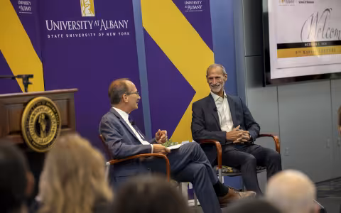 Two men sit in front of a purple backdrop with the words University at Albany on it while speaking before an audience of students and alumni.
