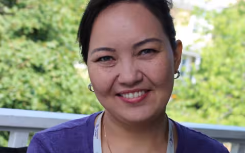 Aizhan smiles at the camera. She is wearing a purple shirt and behind her are green trees.