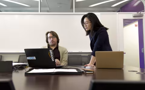 A professor sits at a desk before a computer as a student looks on