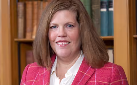 Jessica T. smiles at the camera, wearing a pink blazer and standing in front of a bookcase.