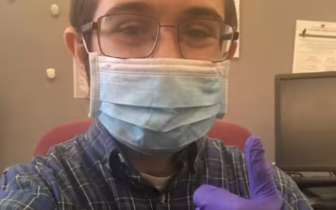 Monroe sits at a desk, his face covered by a mask. He holds his hand, covered by a purple latex glove, in a "thumbs up" position.