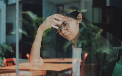 A woman sits at a wooden coffee table in a shop. She has her forehead leaning against her hand, and she appears stressed.