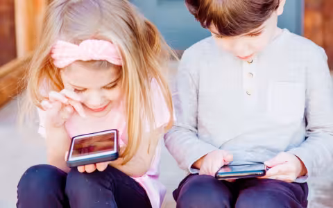 A blond girl in a pink shirt and jeans and a boy in a grey shirt and jeans sit outside on a step, playing on cell phones.