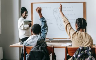 Young students raise their hands as a teacher smiles at the blackboard