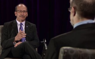 President Rodríguez is seen seated in the left of the frame, speaking to a seated man who appears as a blurry image in the right