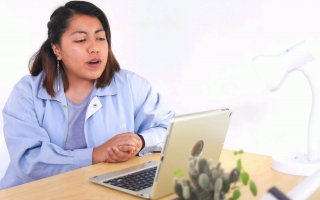 A woman in a blue shirt talks, sitting at a desk in front of a laptop computer