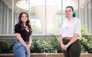 Two young women stand, both with hands clasped in front of them, in front of series of windows