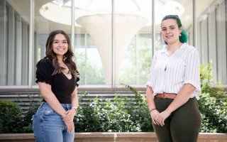 Two young women stand, both with hands clasped in front of them, in front of series of windows
