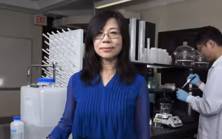 UAlbany Professor of Environmental and Sustainable Engineering Yanna Liang stands in her lab in front of equipement as one of her researchers uses a pipette in the background.