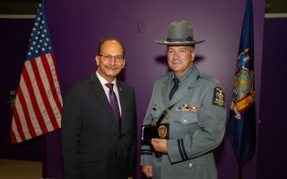 A smiling man in a dark suit and an officer in State Police uniform stand between the U.S. and N.Y. flags